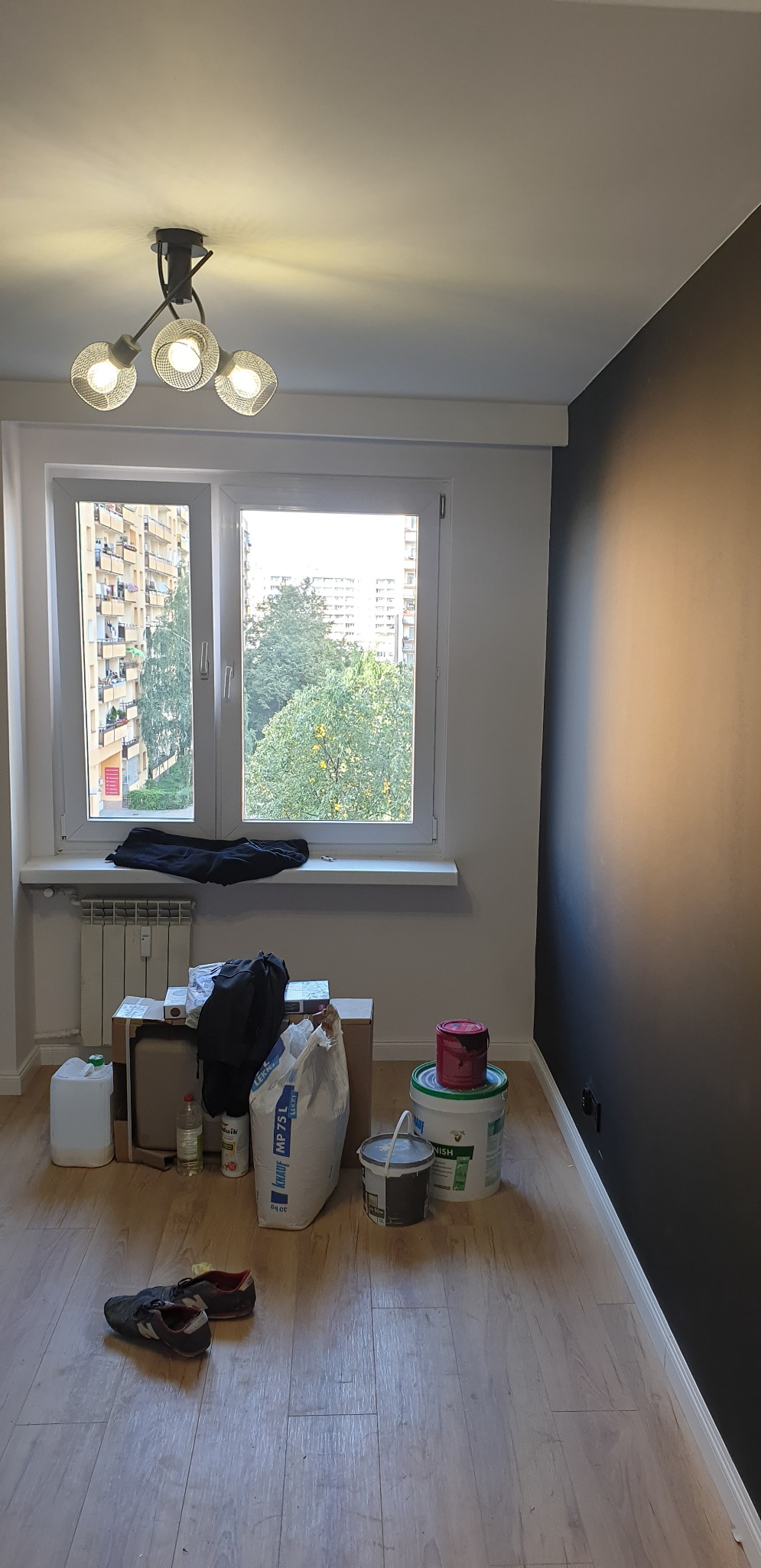 Pokój w trakcie malowania: jedna ściana pomalowana na ciemny kolor, widoczne wiaderka z farbą, torba z gładzią szpachlową Knauf MP 75 L, okno z widokiem na blok, na parapecie leżą ubrania,...