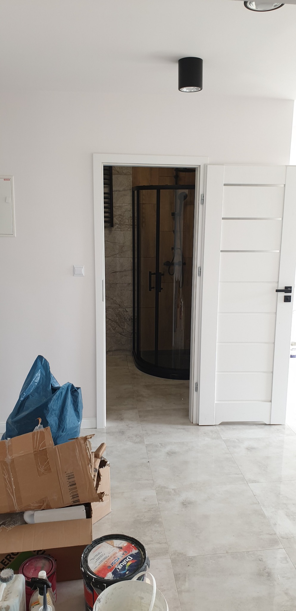 Widok na kabinę prysznicową z czarnymi profilami, widoczną przez otwarte białe drzwi w trakcie prac wykończeniowych, z workami i puszkami farby na pierwszym planie.