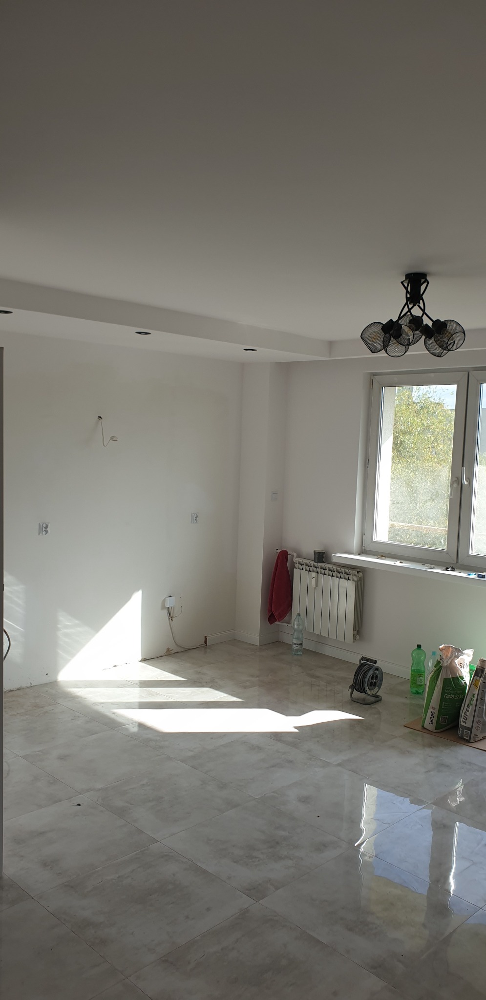 Jasne wnętrze po remoncie z nową, błyszczącą podłogą z płytek, widoczne worki z zaprawą, grzejnik, okno i lampa sufitowa.