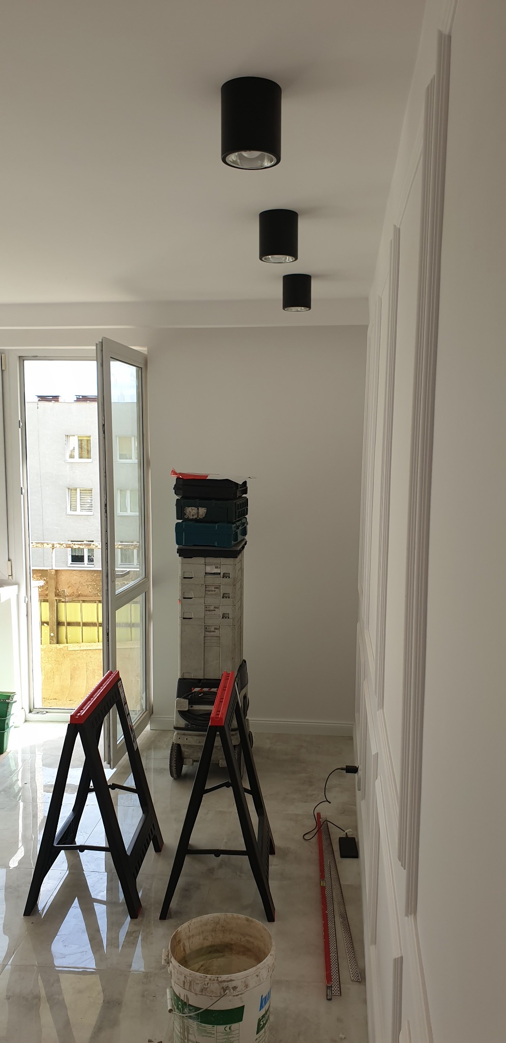 Wnętrze w trakcie prac wykończeniowych: widoczne czarne lampy sufitowe, dekoracyjna sztukateria na ścianie, drabina na kółkach z narzędziami, kozły malarskie i wiadro z zaprawą na marmurowej posadzce.