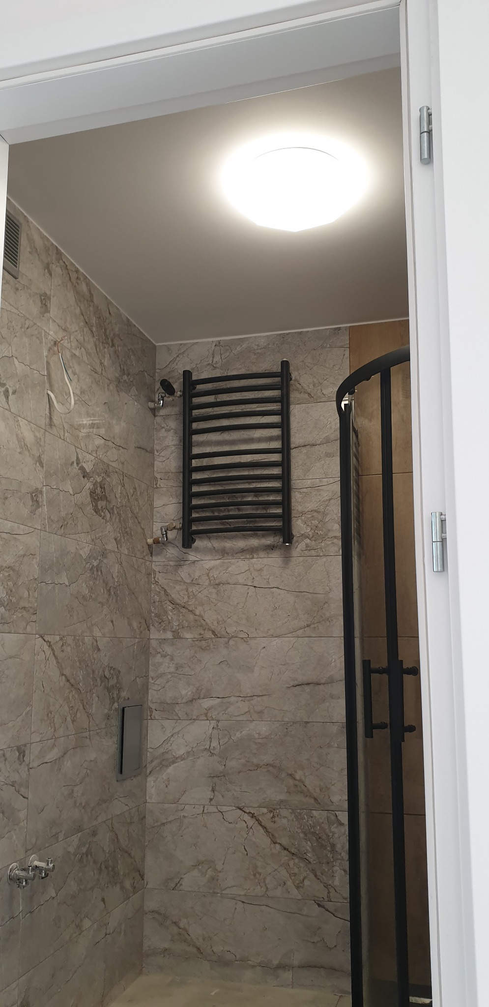 Wykończona łazienka z kabiną prysznicową z czarnymi profilami, grzejnikiem drabinkowym w kolorze czarnym i płytkami imitującymi marmur na ścianach.