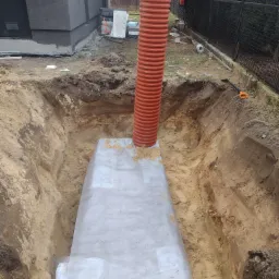 Wykopany rów z pomarańczową rurą karbowaną wystającą pionowo, przykryty szarą folią ochronną, widoczna łopata, obok budynek i ogrodzenie.