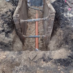 WIZKOP MICHAŁ WIESE - Zabezpieczony wykop z pomarańczową rurą kanalizacyjną w środku, widoczne metalowe szalunki i betonowe kręgi studni w tle.