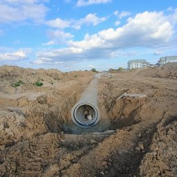 WIZKOP MICHAŁ WIESE - Betonowa rura kanalizacyjna w wykopie ziemnym, widok z góry na wnętrze rury, w tle błękitne niebo z chmurami i budynki mieszkalne.
