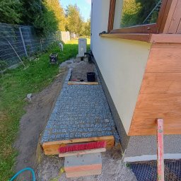 Wiz.kop - Fragment układanej ścieżki z kostki granitowej przy ścianie budynku, widoczne narzędzia, poziomica, szczotka, sznurek traserski i przygotowane podłoże.