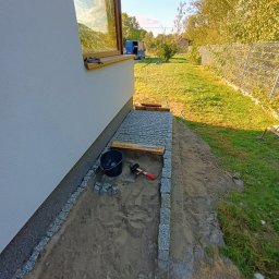 Wiz.kop - Przygotowanie podłoża pod ścieżkę z kostki granitowej przy ścianie budynku, widoczne obrzeża z kostki, piasek, poziomica, młotek, wiadro i częściowo ułożona kostka. W tle trawnik i ogrodzenie.