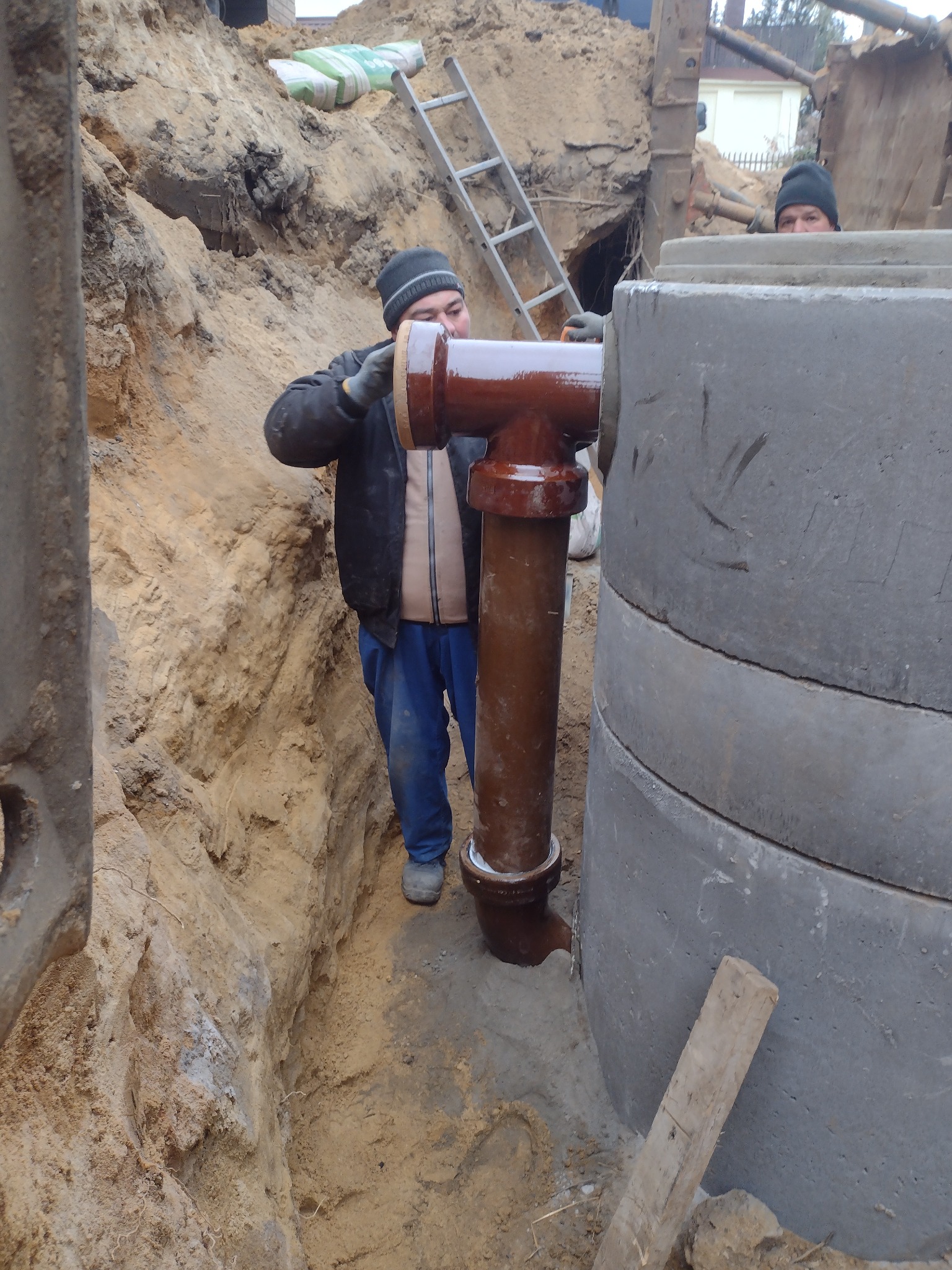 Pracownik w wykopie łączy brązową rurę kanalizacyjną w kształcie litery T z betonowym kręgiem studni; w tle widoczna drabina i worki z materiałami budowlanymi.