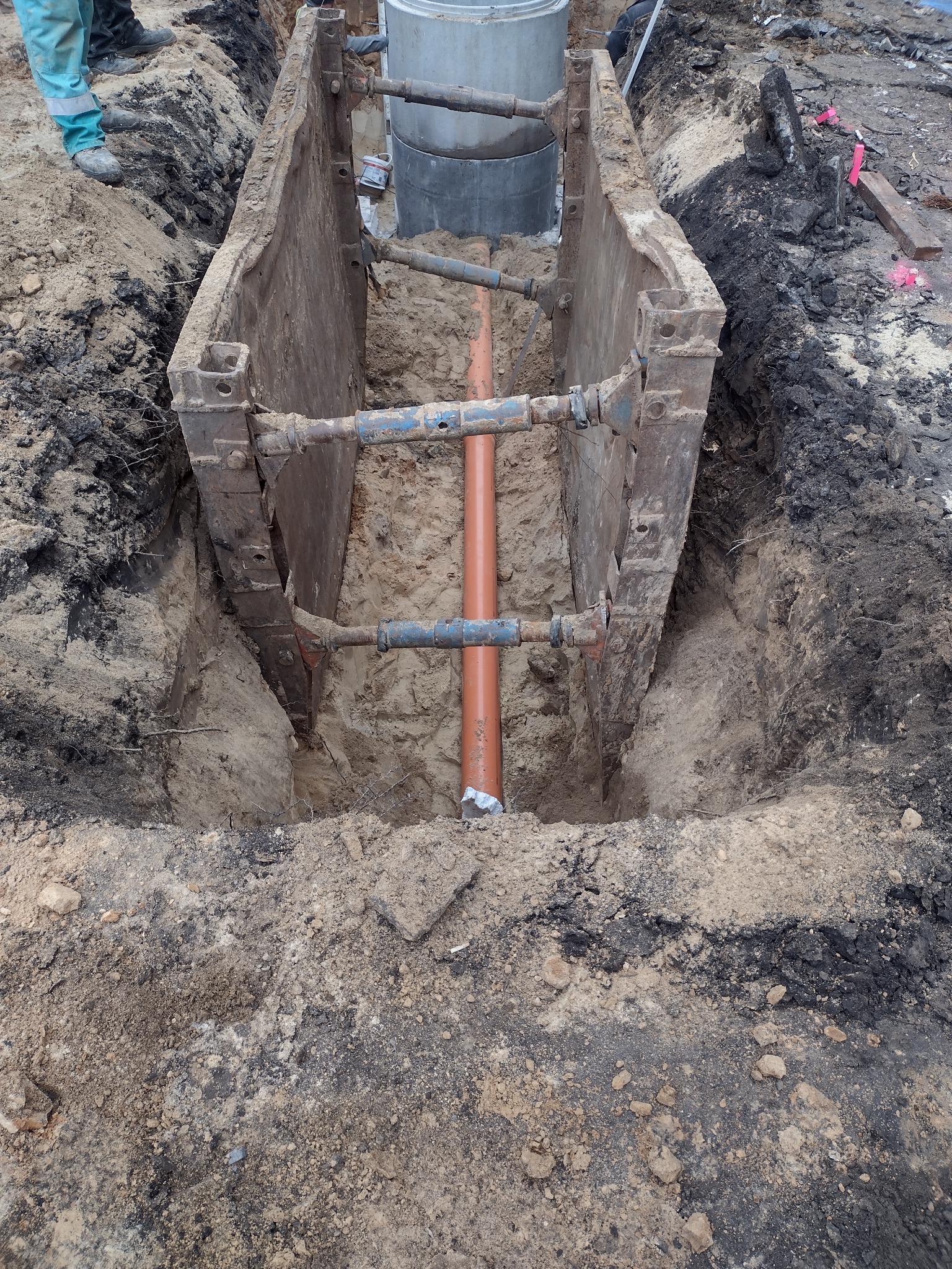 Zabezpieczony wykop z pomarańczową rurą kanalizacyjną w środku, widoczne metalowe szalunki i betonowe kręgi studni w tle.