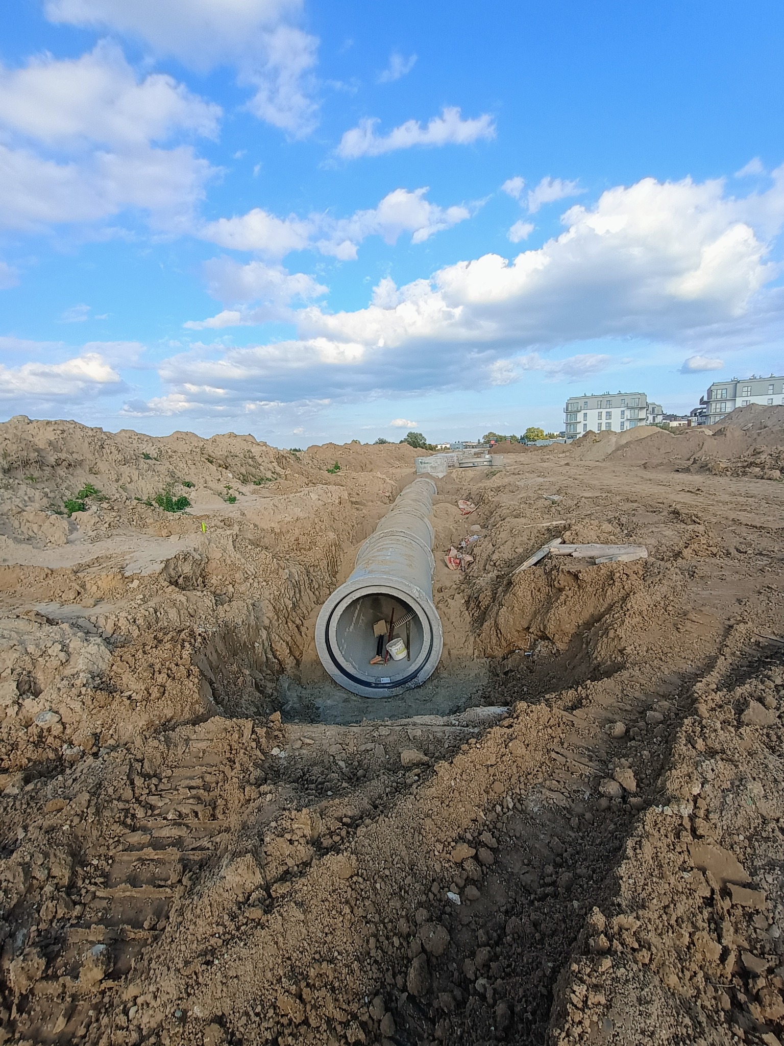 Betonowa rura kanalizacyjna w wykopie ziemnym, widok z góry na wnętrze rury, w tle błękitne niebo z chmurami i budynki mieszkalne.