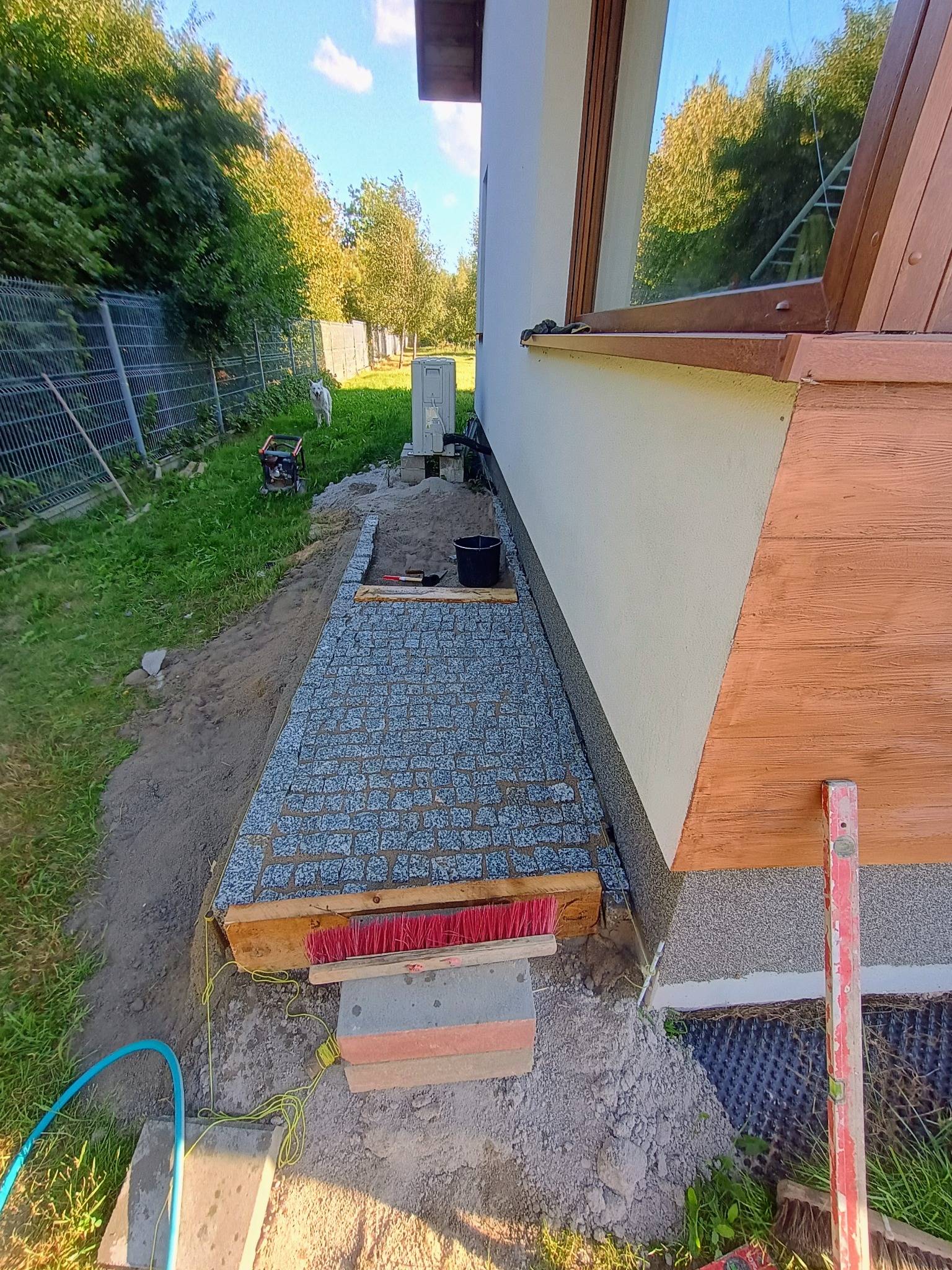 Fragment układanej ścieżki z kostki granitowej przy ścianie budynku, widoczne narzędzia, poziomica, szczotka, sznurek traserski i przygotowane podłoże.