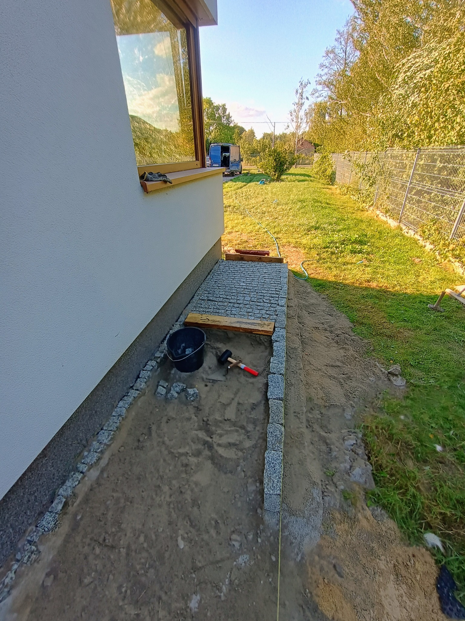 Przygotowanie podłoża pod ścieżkę z kostki granitowej przy ścianie budynku, widoczne obrzeża z kostki, piasek, poziomica, młotek, wiadro i częściowo ułożona kostka. W tle trawnik i ogrodzenie.