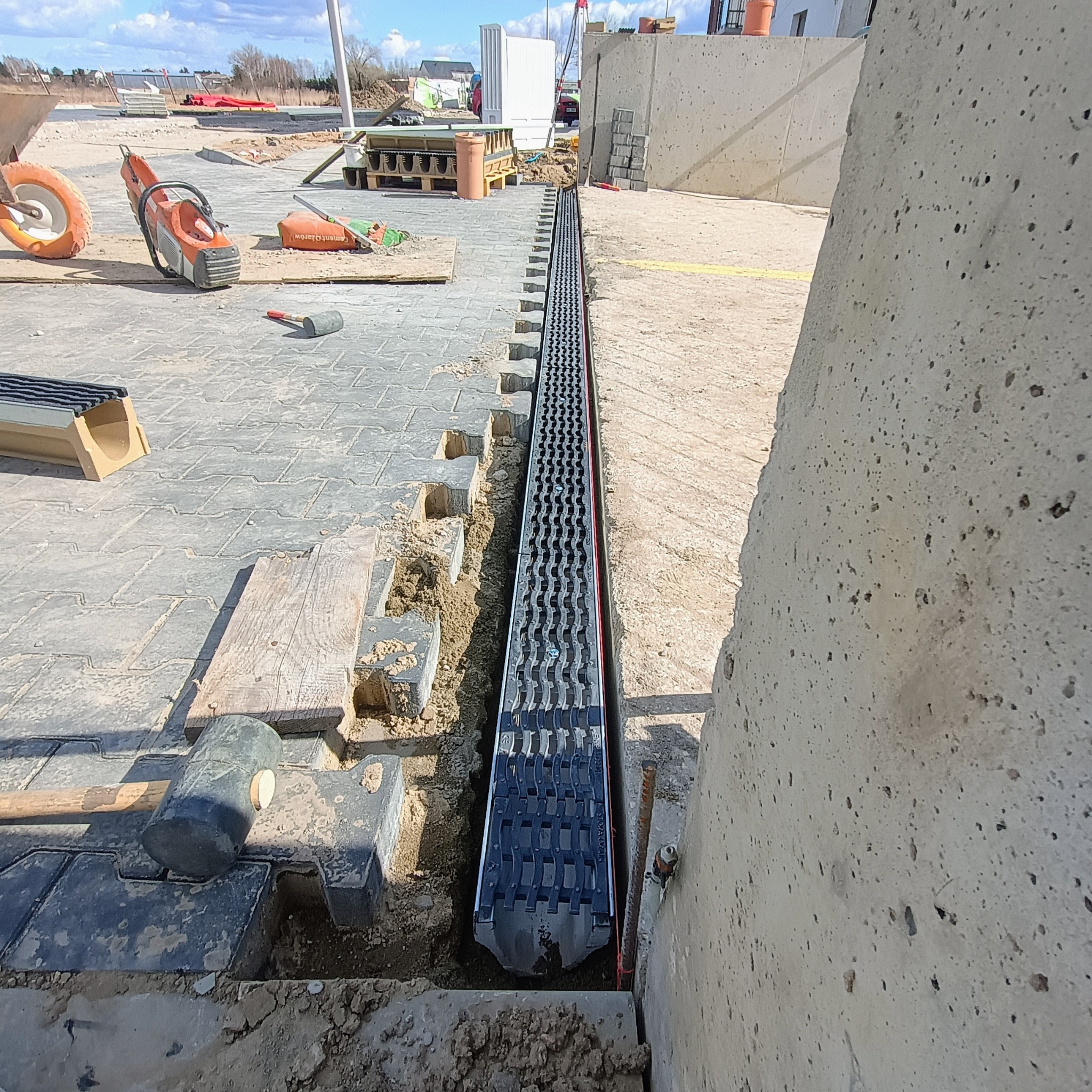 Montaż liniowego systemu odwadniającego z czarną kratką na tle ułożonej kostki brukowej, widoczne narzędzia: gumowy młotek i piła do cięcia kostki.