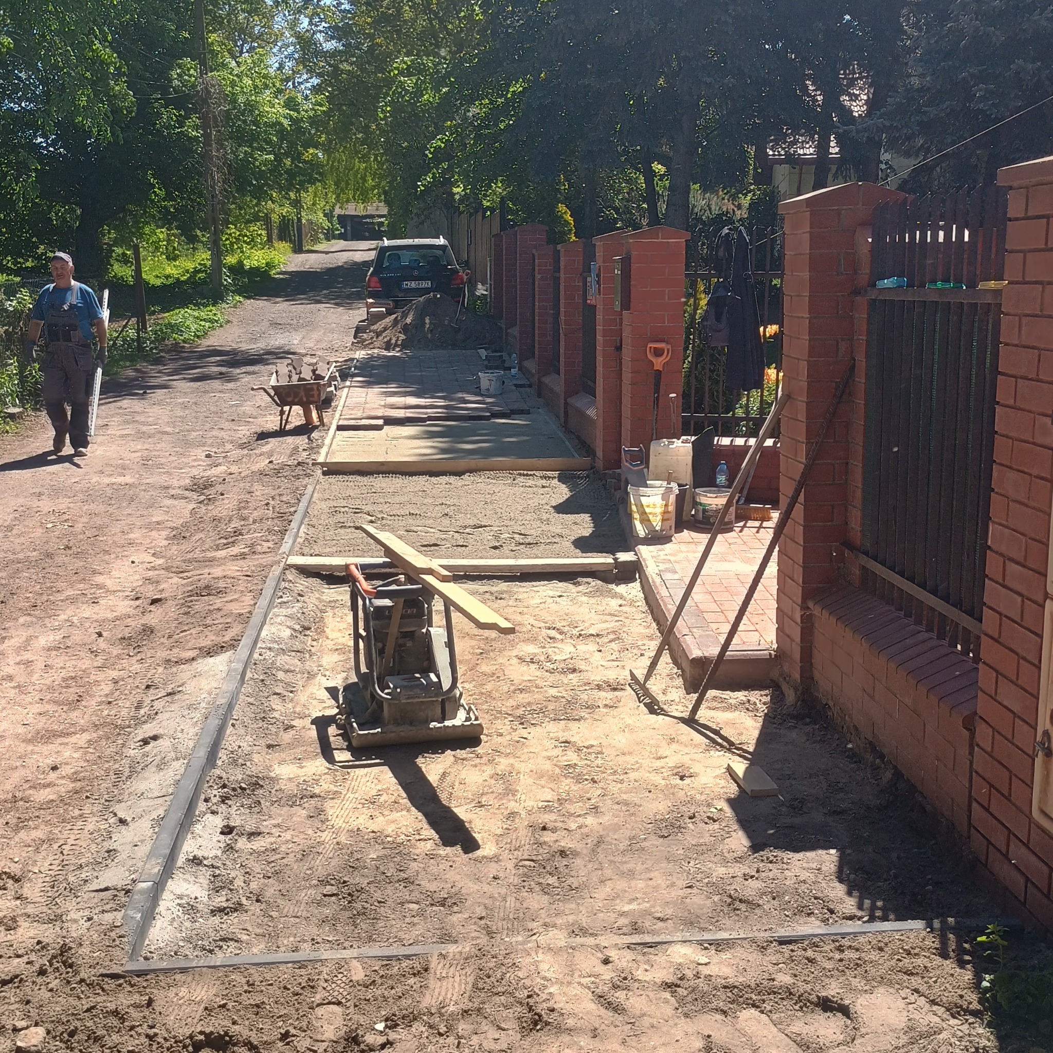 Etap prac brukarskich: ubijanie podłoża pod kostkę brukową zagęszczarką, widoczne obrzeża i częściowo ułożona kostka przy ogrodzeniu z cegły.