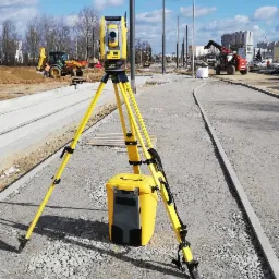 Żółty teodolit na trójnogu i żółta skrzynka na tle budowy drogi z koparką i podnośnikiem w tle, błękitne niebo