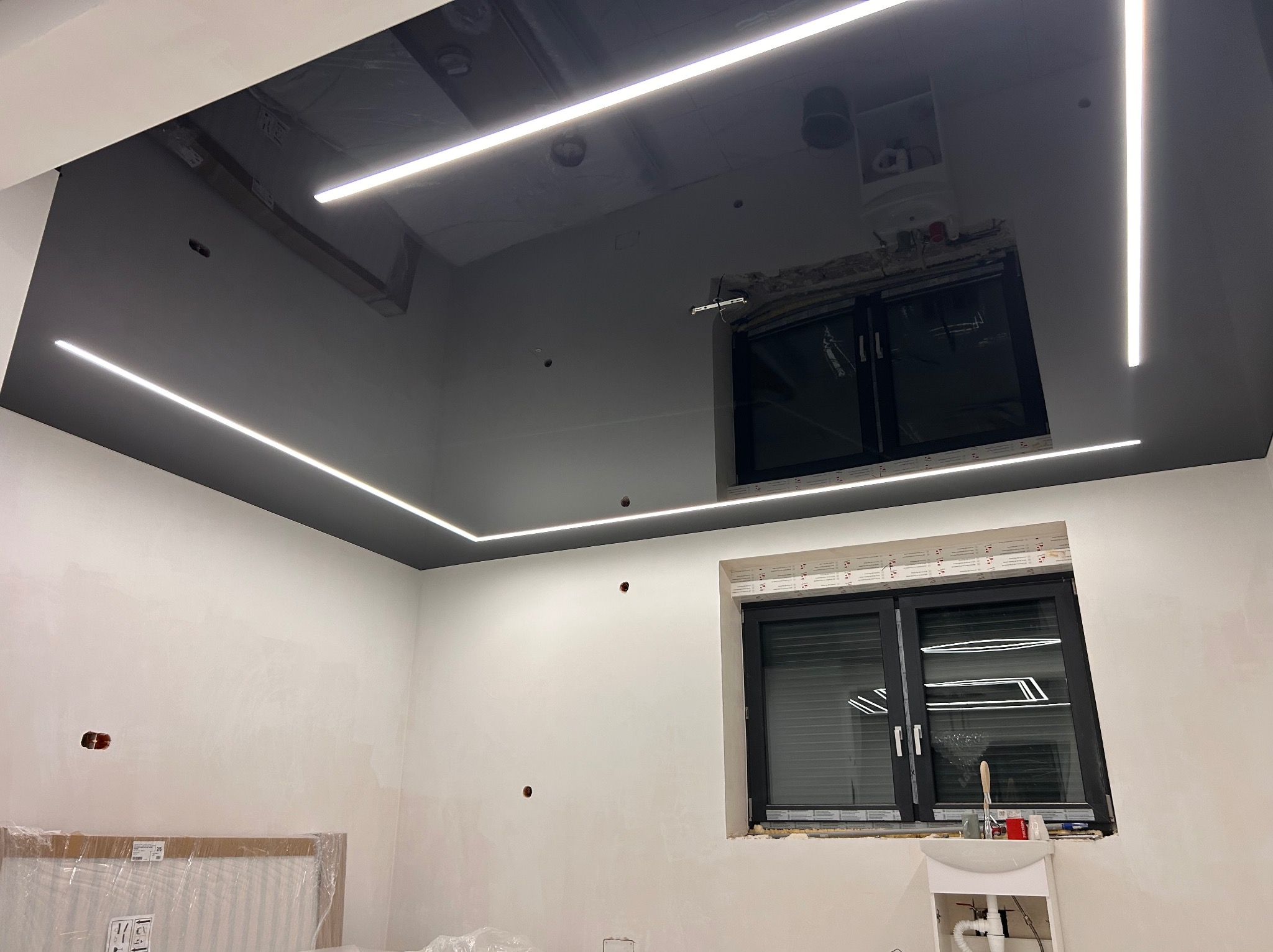 Błyszczący, czarny sufit napinany z liniowym oświetleniem LED w nowo wykończonym wnętrzu. Widoczne okna i ściany przygotowane do malowania.