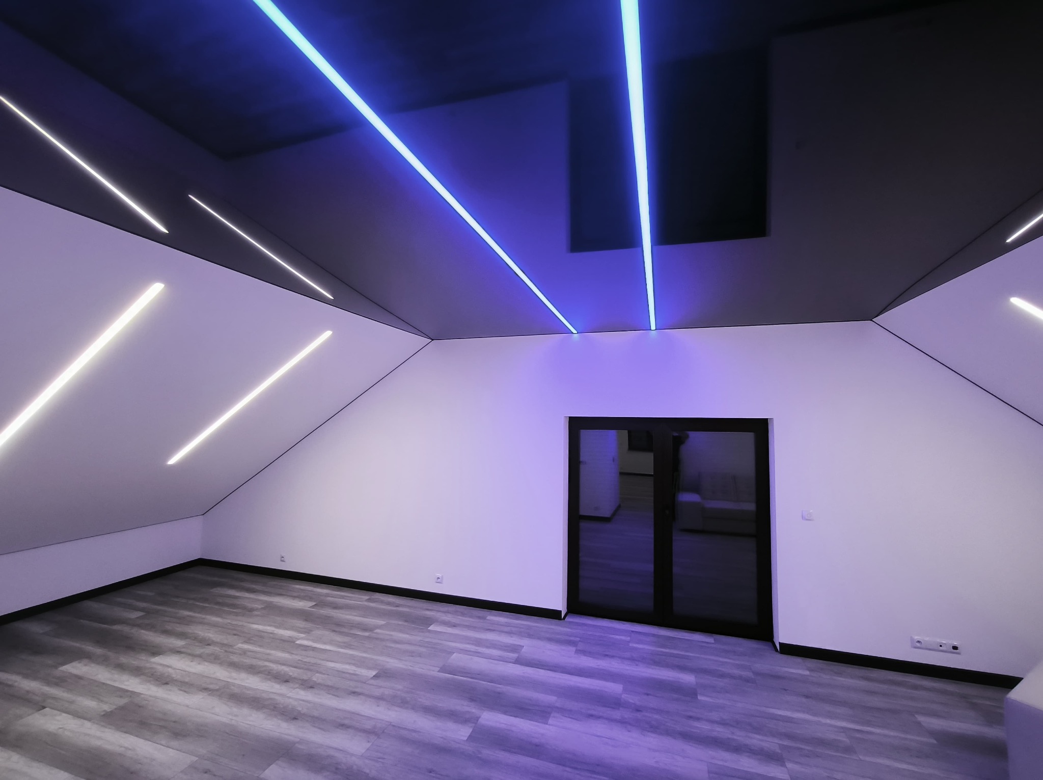 Nowoczesne poddasze z oświetleniem LED: białe ściany, szary parkiet, czarny sufit z wbudowanymi panelami i neonowymi liniami. Minimalistyczny design, widok na dwuskrzydłowe drzwi.