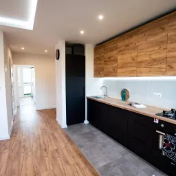 Nowoczesna kuchnia z czarnymi szafkami dolnymi i drewnianymi szafkami górnymi, podświetlona listwą LED pod szafkami, z szarą podłogą i drewnianą podłogą w przedpokoju.