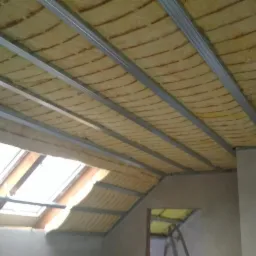 Ocieplone wełną mineralną poddasze z widoczną konstrukcją metalową pod płyty gipsowe, okno dachowe w tle.