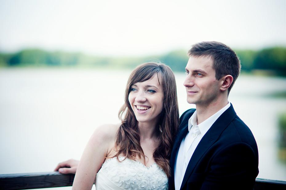 Uśmiechnięta para młoda, panna młoda w białej sukni z koronkowym gorsetem, pan młody w czarnym garniturze i białej koszuli, stojąca na drewnianym pomoście z widokiem na jezioro i zielone drzewa w tle.