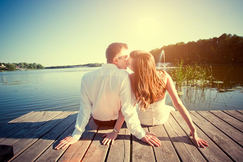 Para młoda siedzi na drewnianym pomoście nad jeziorem, w tle fontanna wodna i las, pocałunek w promieniach słońca.