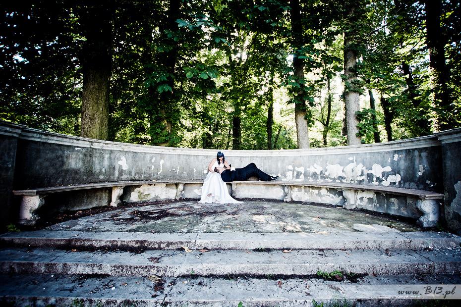 Panna młoda w białej sukni i welonie siedzi na kamiennej ławce, obok leży pan młody w czarnym garniturze, w tle park z drzewami.
