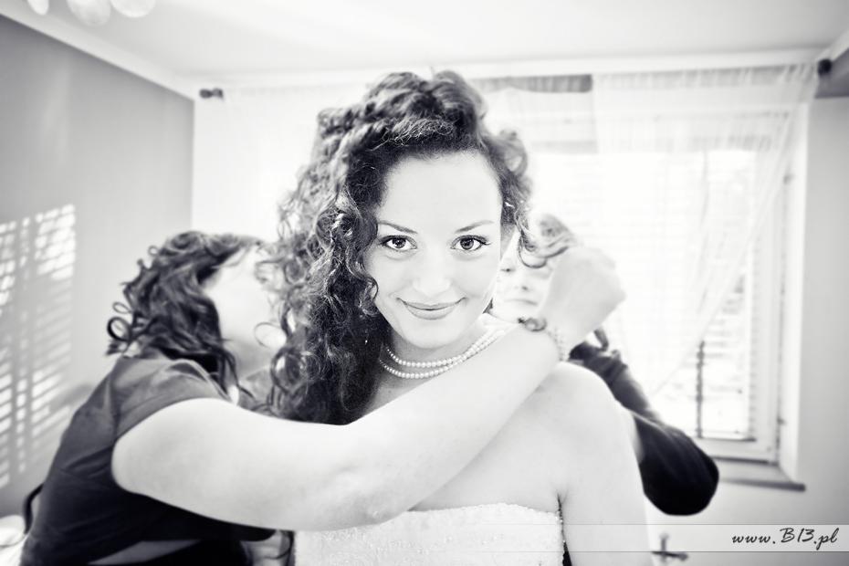 Czarno-białe zdjęcie uśmiechniętej panny młodej z kręconymi włosami, podczas przygotowań do ślubu w Bydgoszczy, z pomocą osób trzecich dopinających jej naszyjnik z pereł.