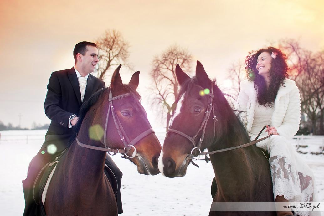 Para młoda w strojach ślubnych siedzi na koniach w zimowej scenerii, konie stoją naprzeciwko siebie.