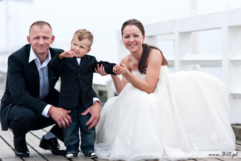 Uśmiechnięta para młoda z synkiem w garniturze pozuje na drewnianym pomoście, on kuca, ona siedzi w sukni ślubnej, w tle białe barierki.