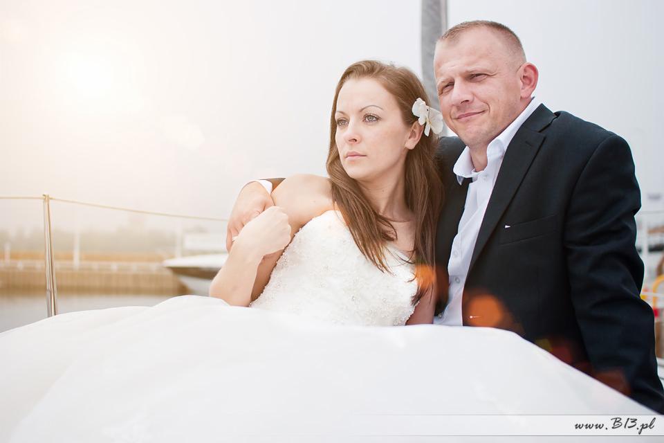 Para młoda na jachcie: panna młoda w białej sukni z kwiatem we włosach, pan młody w czarnym garniturze, oboje patrzą w dal.