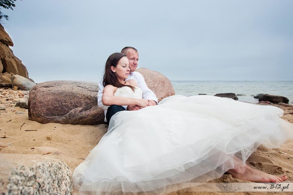 Para młoda siedzi na piasku przy morzu, panna młoda w sukni z koronkowymi aplikacjami, pan młody obejmuje ją ramionami, w tle kamienie i spokojna woda.