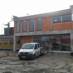 Budynek w trakcie montażu okien PCV, widoczne surowe ściany z cegły, zamontowane okna w czarnych ramach oraz samochód dostawczy na placu budowy