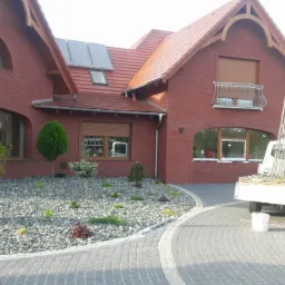 Dom z czerwonej cegły z brązowymi roletami zewnętrznymi i oknami, na podjeździe zaparkowany biały samochód dostawczy z elementami montażowymi.