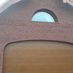 Garaż z cegły klinkierowej z drewnianą bramą segmentową i półokrągłym oknem na szczycie