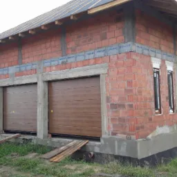 Surowy budynek w trakcie budowy z zamontowanymi brązowymi segmentowymi bramami garażowymi i oknami z ciemnymi ramami, widoczne podpory z desek przed bramami.