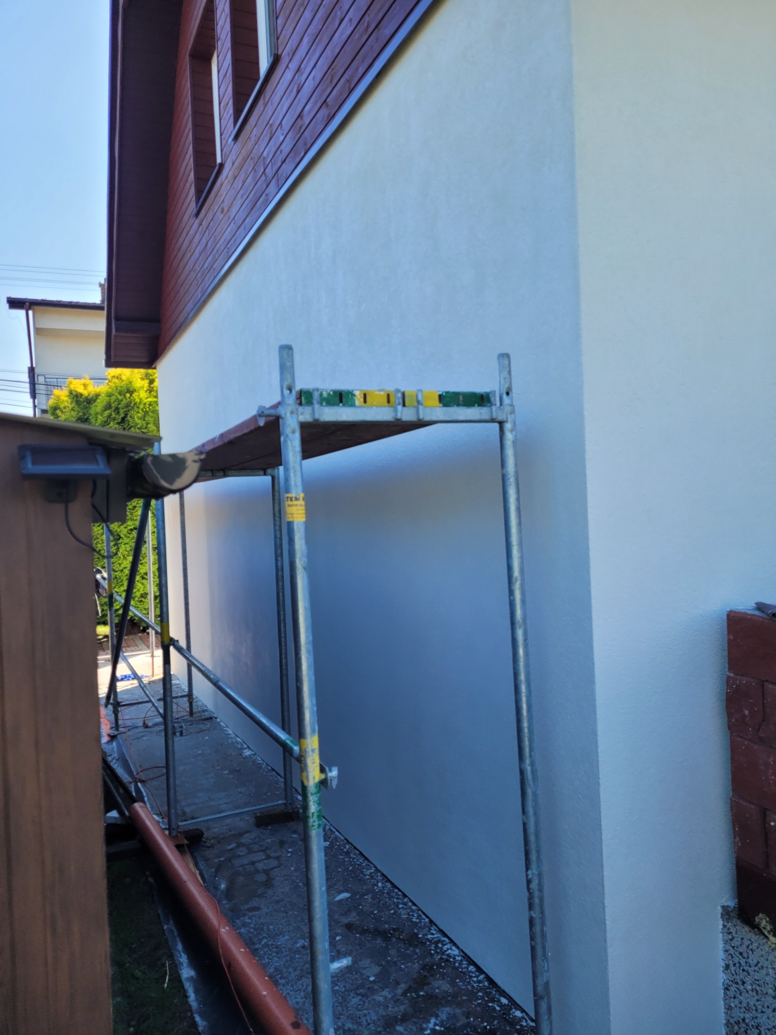 Świeżo pomalowana, jasna elewacja domu jednorodzinnego z widocznym rusztowaniem i brązowym orynnowaniem, fragment drewnianej podbitki dachowej.