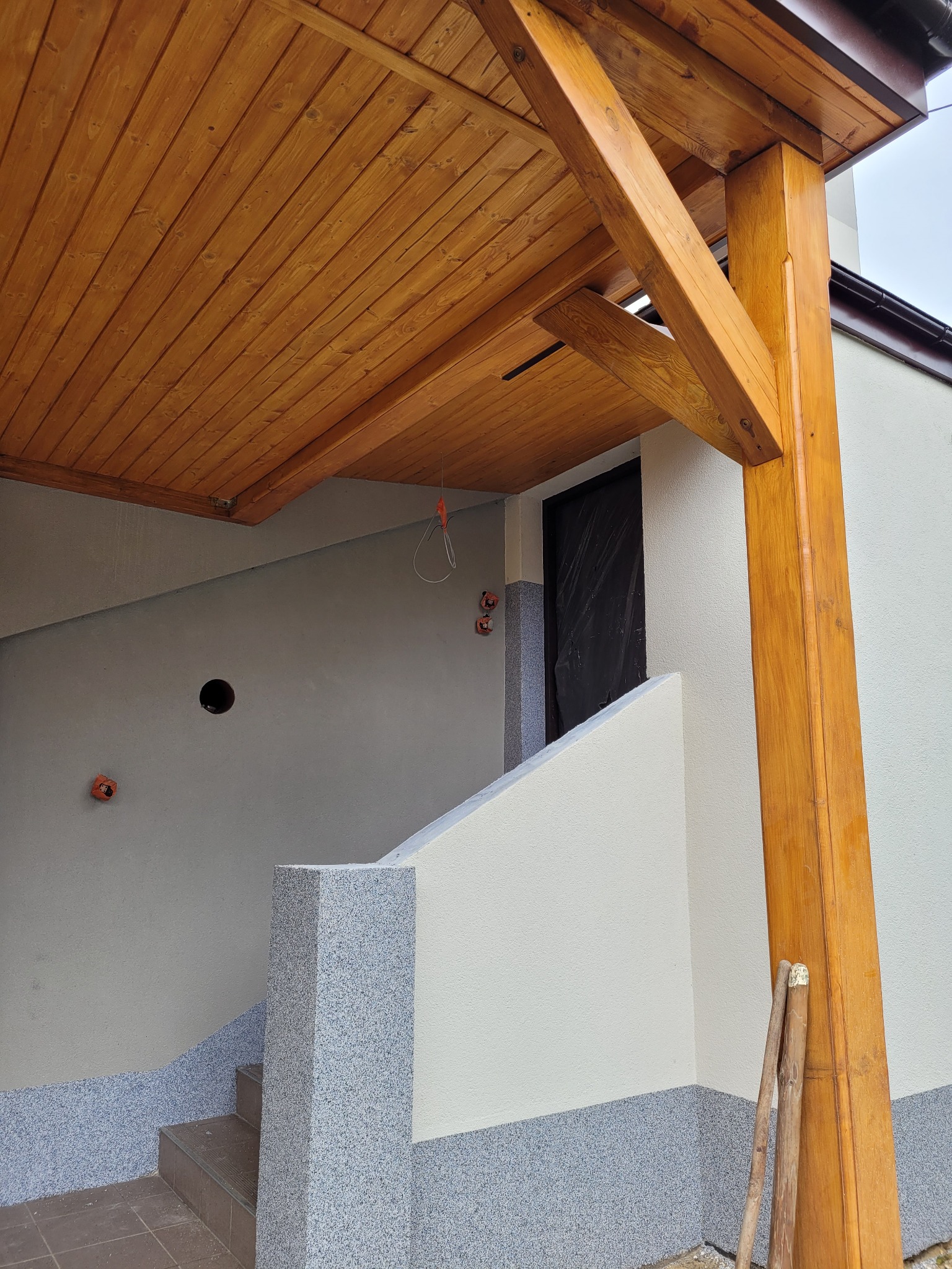 Wejście do budynku z drewnianym zadaszeniem i słupem, widoczne schody z szarą posadzką i fragment elewacji z widocznymi elementami instalacji elektrycznej.