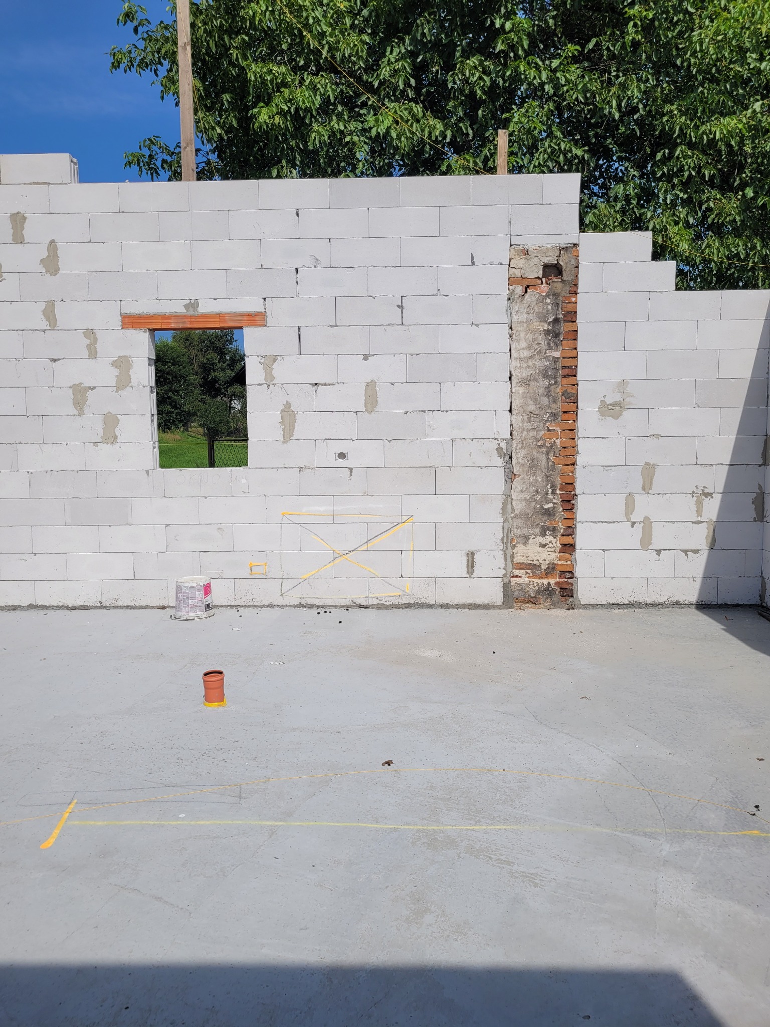 Wznoszenie ściany z bloczków betonu komórkowego z otworem okiennym i fragmentem starej ceglanej ściany, widoczne żółte oznaczenia na posadzce oraz puszka z farbą.