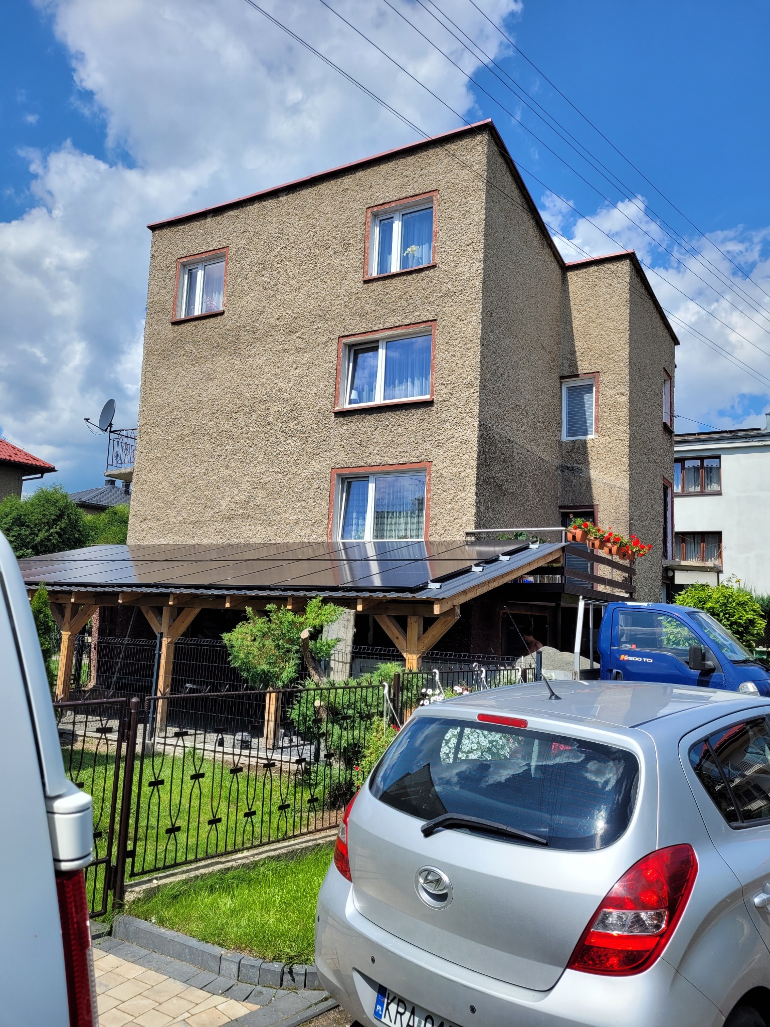 Trzykondygnacyjny dom z elewacją o fakturze baranka, widoczny z poziomu ulicy, z zadaszeniem nad tarasem pokrytym panelami fotowoltaicznymi, z zaparkowanymi samochodami na pierwszym planie.