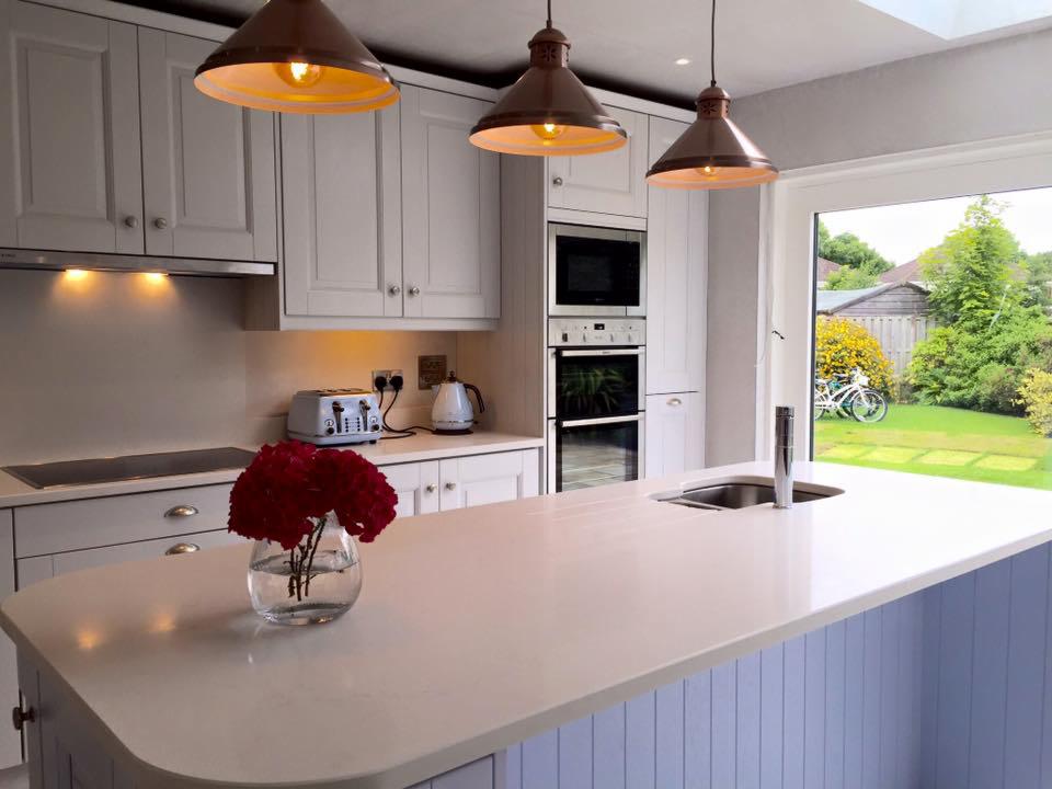 Jasna kuchnia z wyspą i widokiem na ogród, trzy lampy wiszące nad blatem, wazon z czerwonymi kwiatami na pierwszym planie.