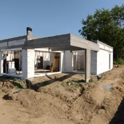 Surowy stan domu z białych pustaków, widoczne zbrojenia i betonowe nadproża, teren wokół w trakcie prac ziemnych, błękitne niebo w tle.