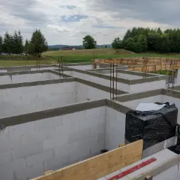 Fundamenty z bloczków betonowych, częściowo zaszalowane drewnem, z wystającymi prętami zbrojeniowymi, na tle zielonego krajobrazu z drzewami i pochmurnym niebem.