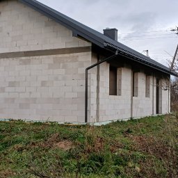 Usługi budowlane EKO-DOMY Tomasz Czuba - Surowy, parterowy dom z białych bloczków, z czarnym dachem i rynnami, w trakcie budowy. Widoczne otwory okienne i fragmenty instalacji. Tło: pochmurne niebo, krzewy.