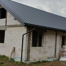 Usługi budowlane EKO-DOMY Tomasz Czuba - Niedokończony dom z białych bloczków, z szarym dachem z blachy. Widoczne niezabezpieczone otwory okienne i drzwiowe. Na ziemi leżą materiały budowlane.