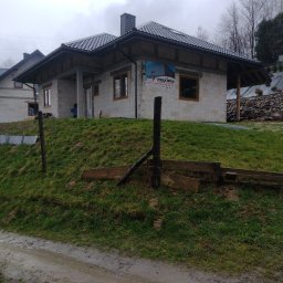 Usługi budowlane EKO-DOMY Tomasz Czuba - Surowy dom z białego pustaka na wzgórzu, z widocznymi oknami i dachem z dachówki. Na ścianie baner reklamowy. Teren błotnisty, pochmurne niebo.