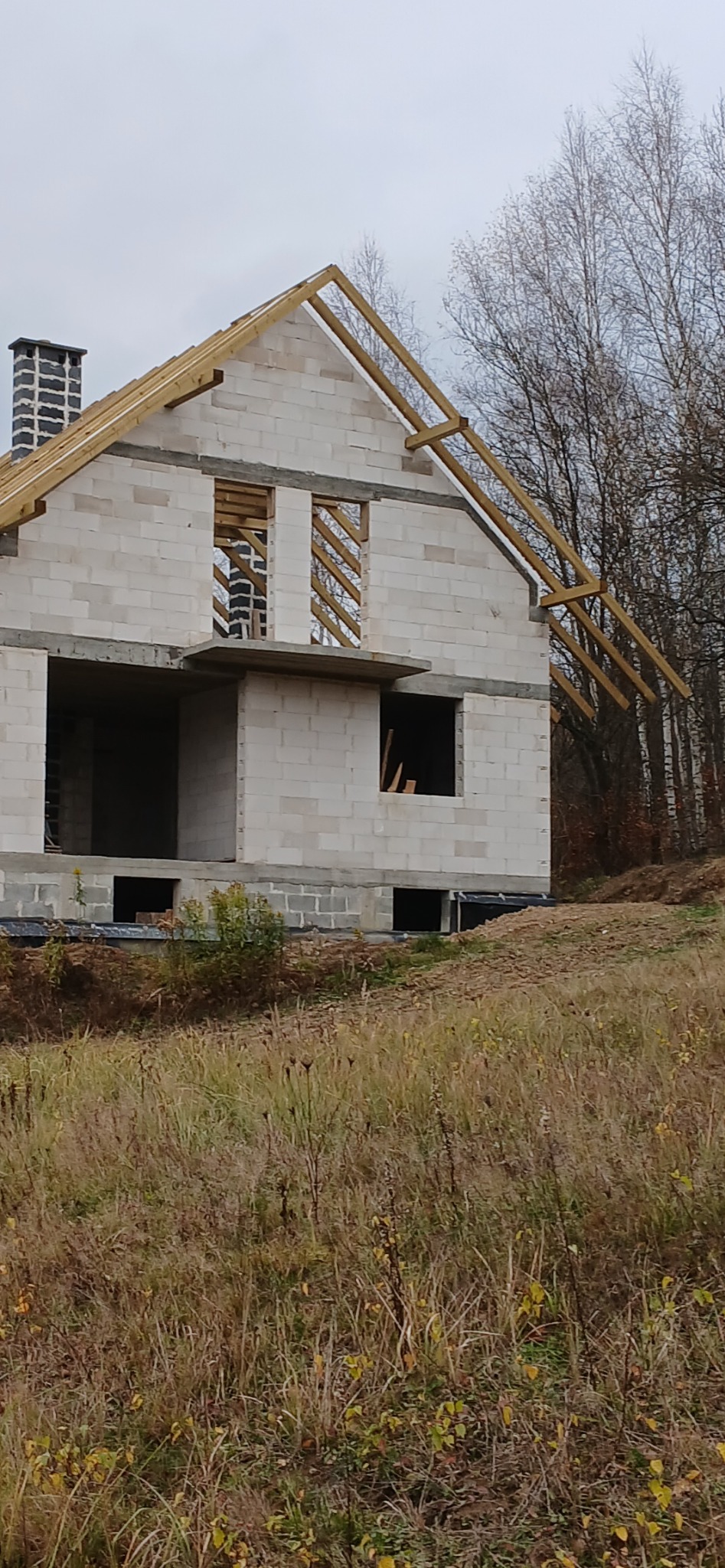 Surowy stan domu z białego betonu komórkowego w trakcie budowy, z widoczną konstrukcją dachu i kominem. Tło stanowią drzewa bez liści i trawa.