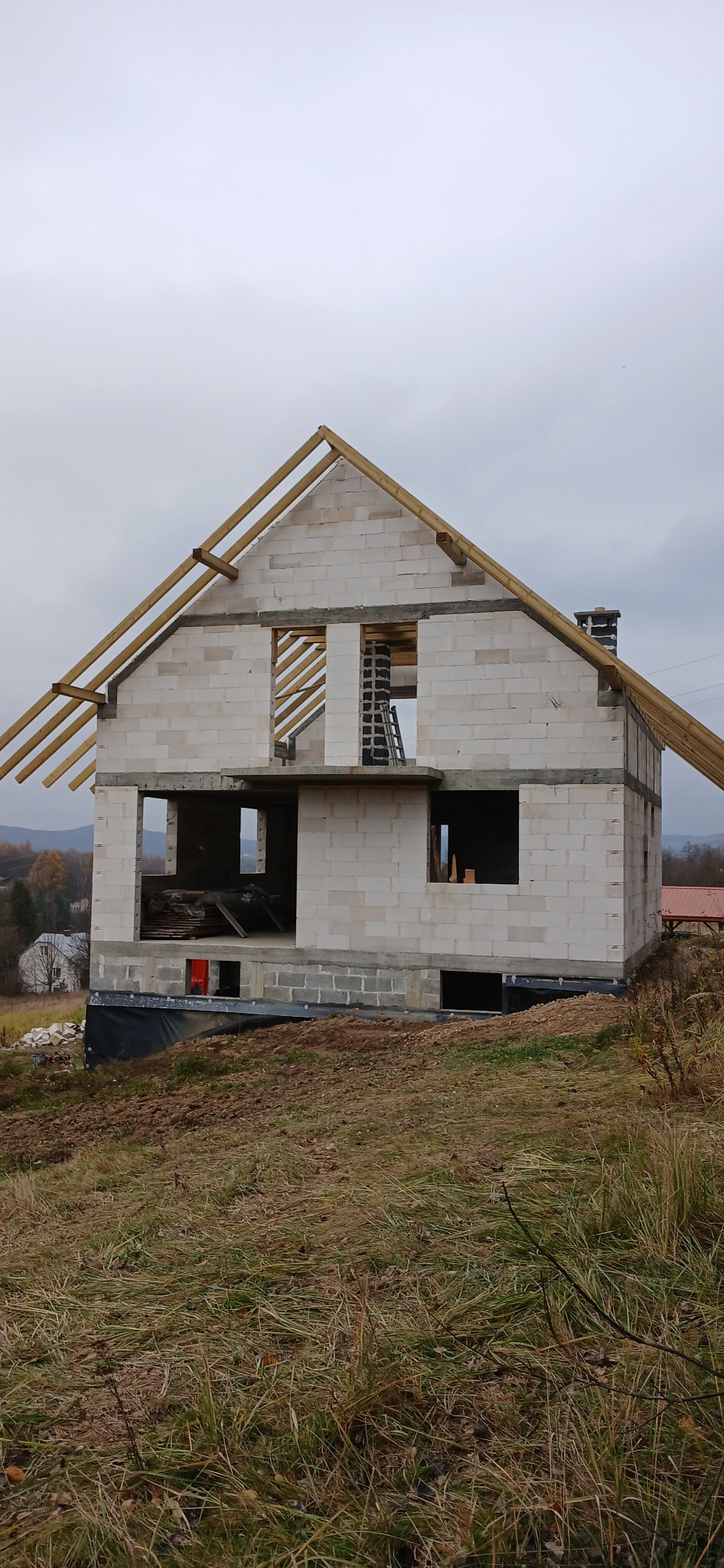 Niedokończony dom z białego betonu komórkowego z drewnianą konstrukcją dachu na tle pochmurnego nieba i pagórkowatego krajobrazu. Widoczna drabina i materiały budowlane.