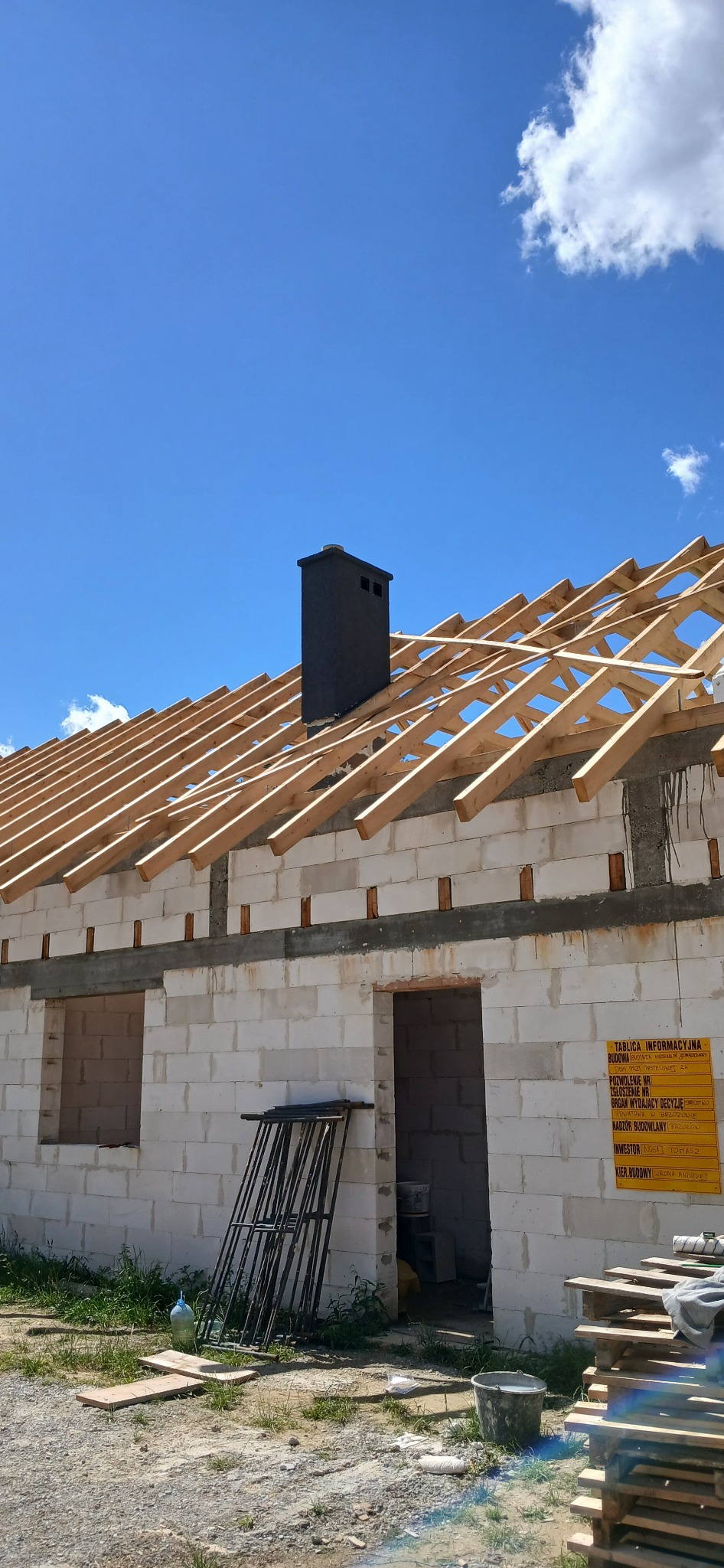 Budowa domu w trakcie: białe ściany z pustaków, drewniana konstrukcja dachu, czarny komin. Tablica informacyjna budowy po prawej stronie. Słoneczny dzień.