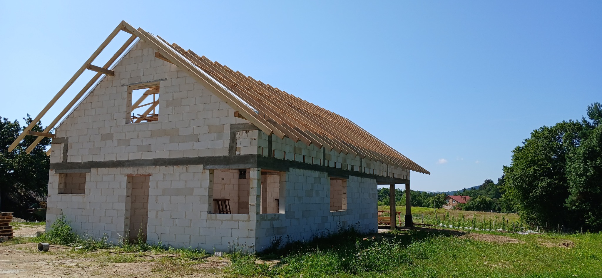 Budowa domu z białych bloczków: ściany wymurowane, wstawione drewniane elementy dachu, widoczne otwory okienne i drzwiowe. W tle zieleń i błękitne niebo.