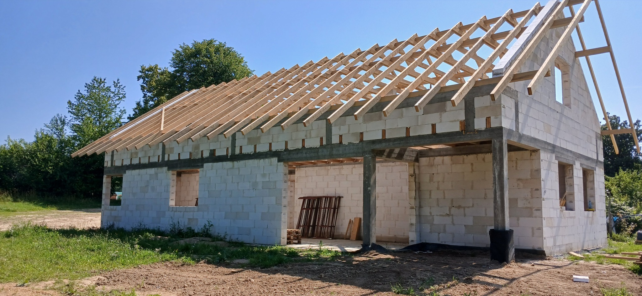 Budowa domu jednorodzinnego w stanie surowym otwartym: białe ściany z betonu komórkowego, drewniana konstrukcja dachu, widoczne zbrojenia. Słoneczny dzień.