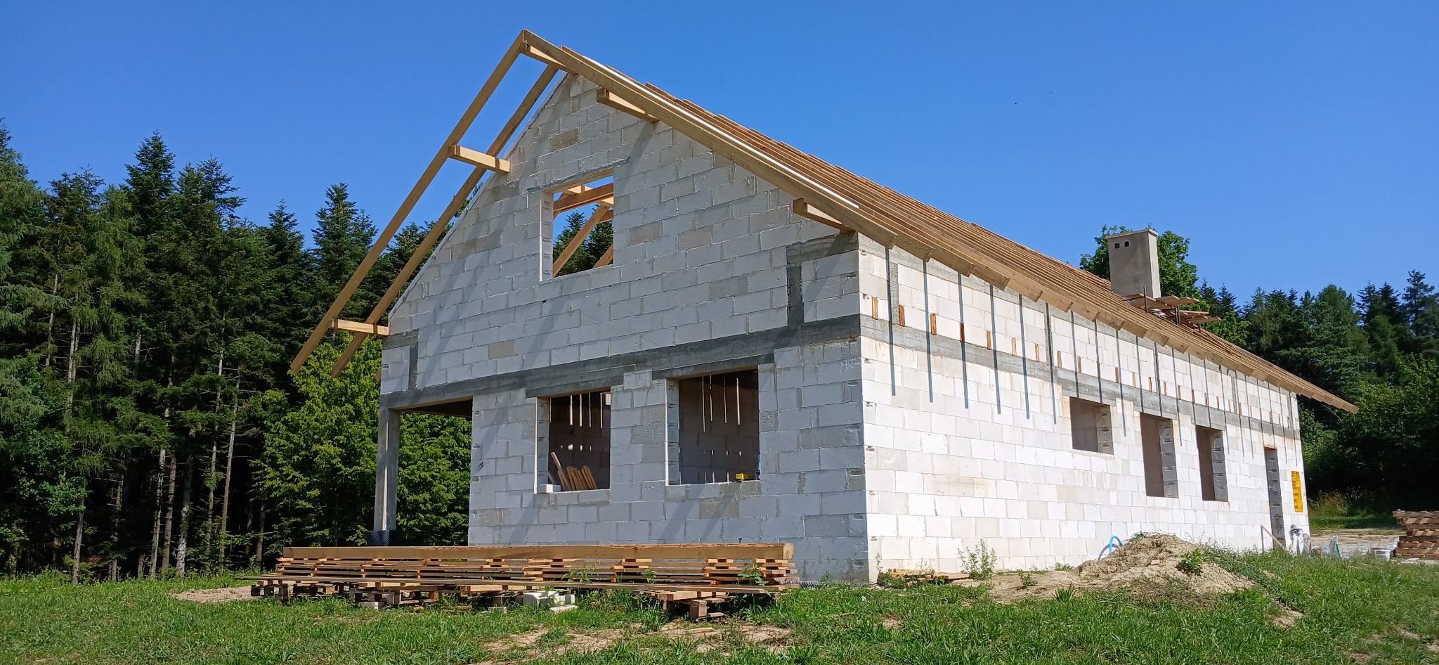 Surowy stan domu z białego betonu komórkowego w trakcie budowy, widoczne drewniane elementy konstrukcji dachu i okna. W tle las iglasty i błękitne niebo.